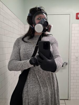 Mirror selfie in a Target restroom, showing Jennifer in a gray skater dress with full-face respirator, gloves, and a really bad makeup job.