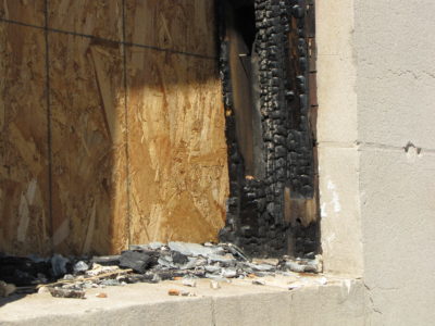 Charred frame and broken glass around the left window on the first floor, next to the main entrance.